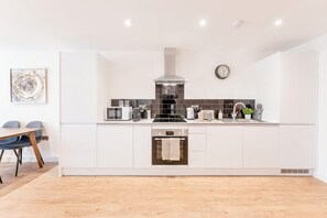 A modern kitchen with white cabinetry, a stainless steel range hood, a built-in oven, and a dining area with a table and chairs. - Spacious & Bright 2 Bed Burton-on-Trent Apartment (Burton upon Trent)