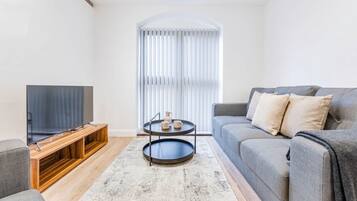 A modern living room with a flat-screen TV on a wooden stand, a grey sofa with cushions, a coffee table with cups, and a large window with blinds.