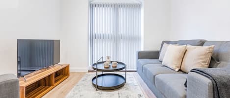 Uma sala de estar moderna com televisão de tela plana em um suporte de madeira, sofá cinza com almofadas, mesa de centro com xícaras e uma janela grande com persianas.