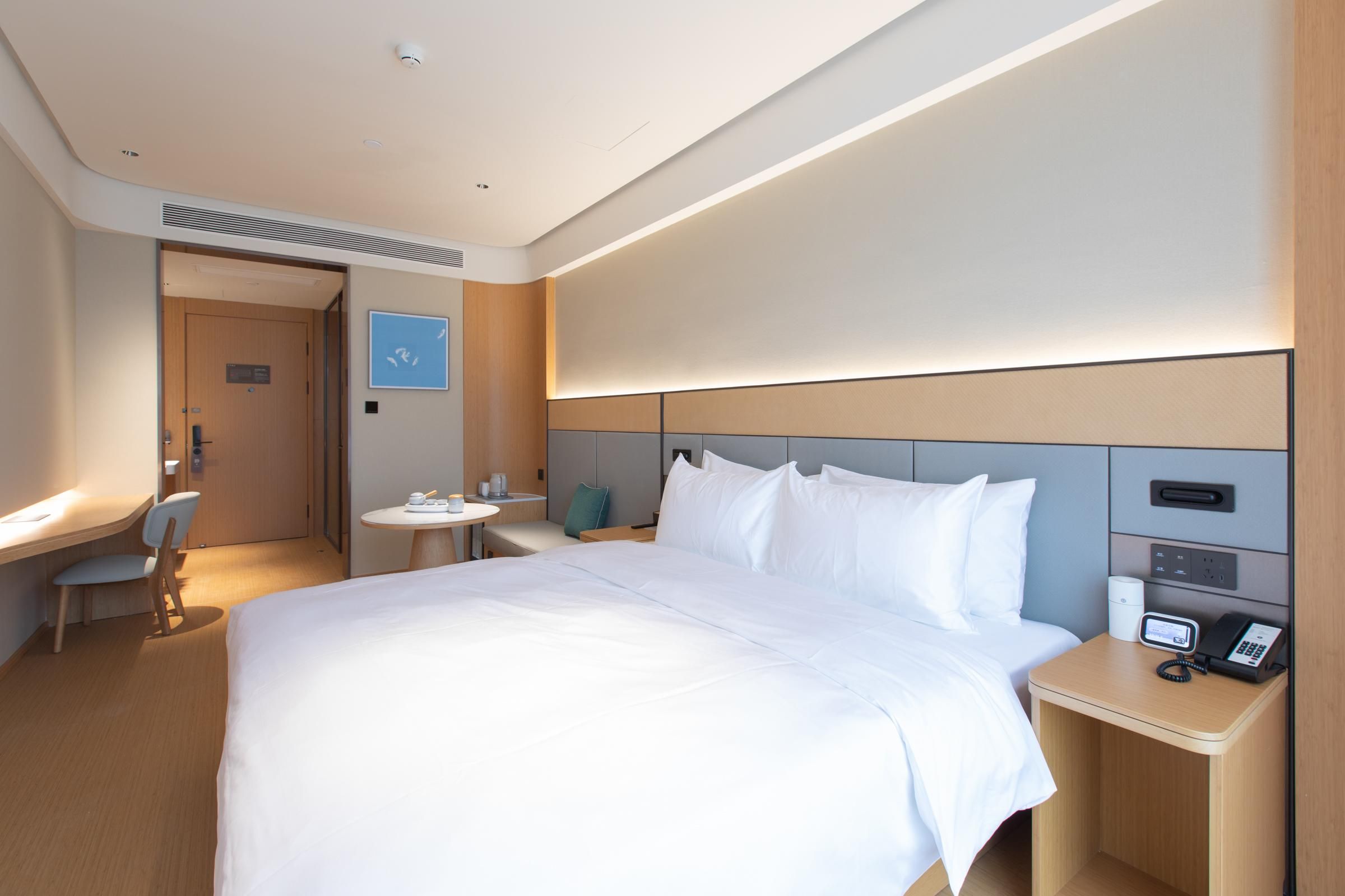A hotel room with a large bed, bedside table with a phone, a small desk with a chair, and a round table with a chair.
