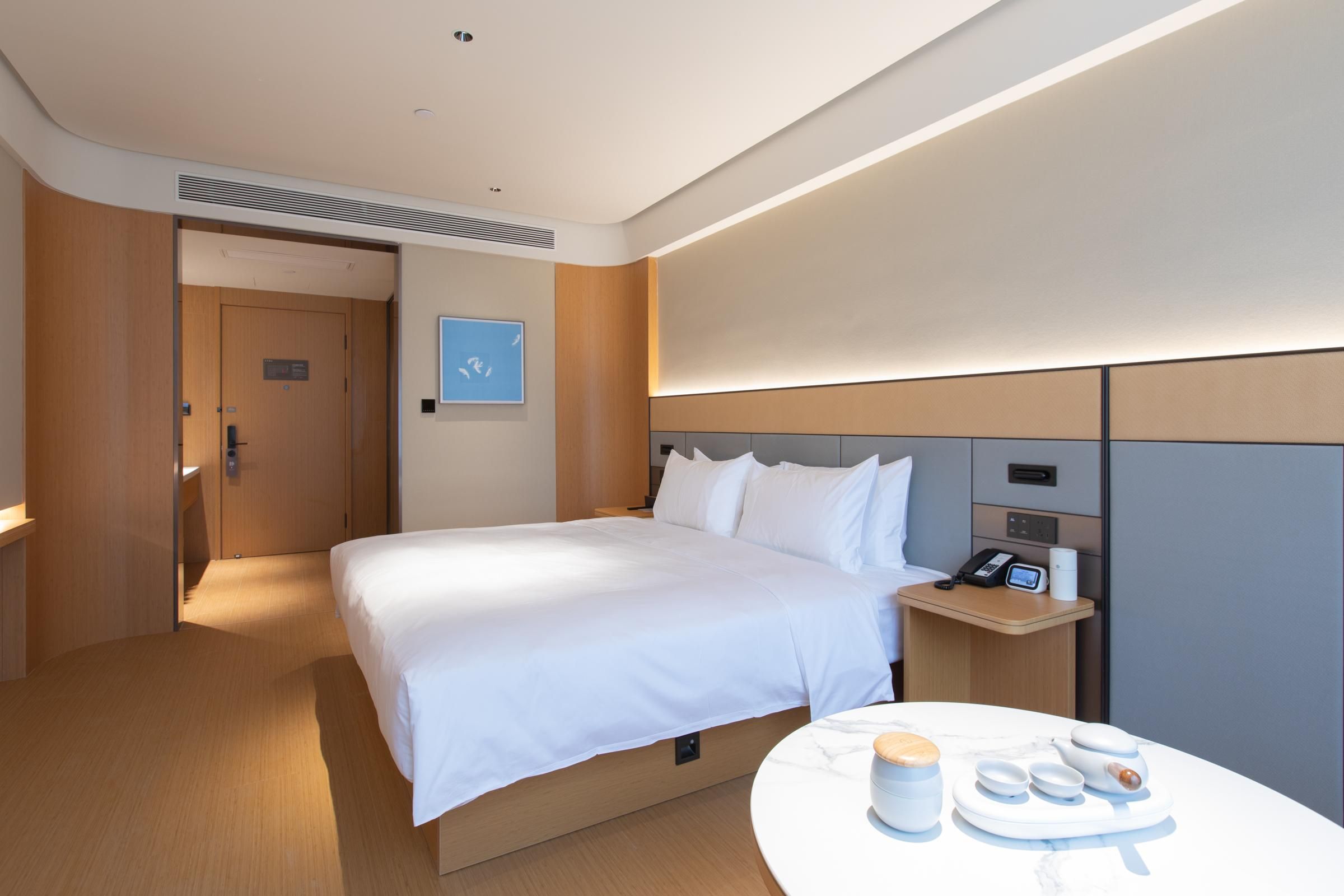 Una habitación de hotel moderna con una cama bien tendida, una mesita de noche con un teléfono y una lámpara pequeña, y una mesa redonda con una bandeja de alimentos para el desayuno.