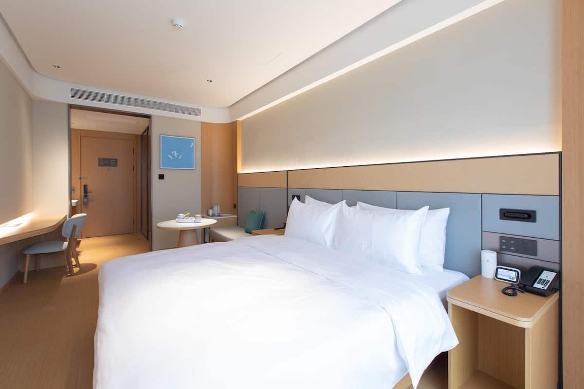 A hotel room with a large bed, bedside table with a phone, a small desk with a chair, and a round table with a chair.