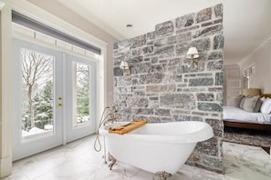 A bathroom with a freestanding bathtub, stone wall, and a view through a glass door. - Badger's Sett - Estate on point with hot tub (Battersea)