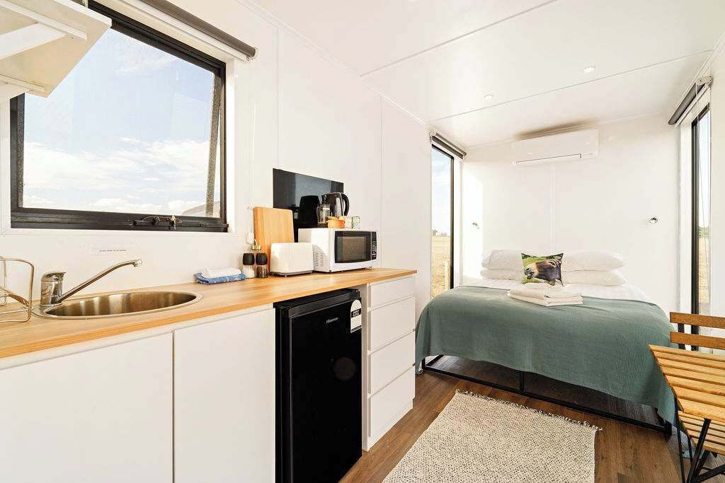 A compact hotel room with a kitchenette, a bed with a green bedspread, and a view through the window.