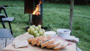 A picnic setup with a variety of cheeses, crackers, and sliced meats on a wooden table, with a grill in the background.