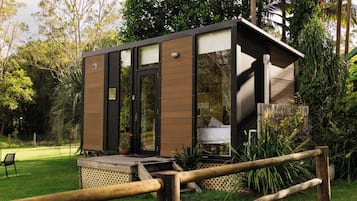 Une petite structure moderne, dotée d’une porte et de fenêtres en verre, entourée d’une végétation luxuriante et d’une clôture en bois.