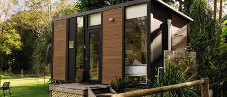 A small, modern structure with a glass door and windows, surrounded by lush greenery and a wooden fence.