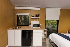 A compact bedroom with a built-in kitchenette, a bed, and a view of greenery.
