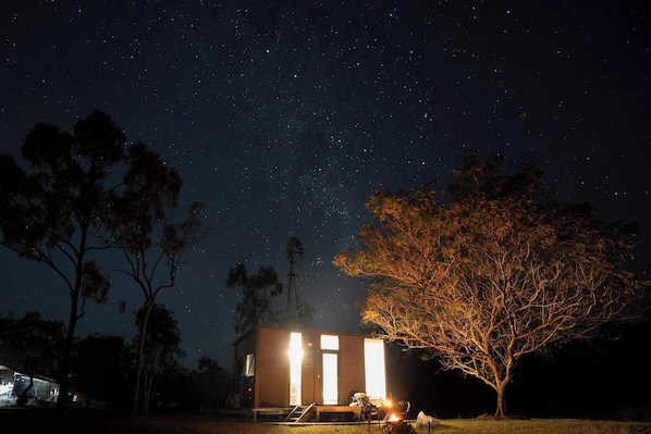 Una cabaña pequeña con un cielo estrellado, un árbol y un molino de viento al fondo.