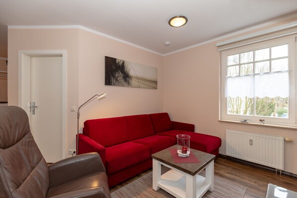 A living room with a red sofa, a brown leather chair, a small white table with a glass vase, a floor lamp, and a window with a view of trees. - Ostsee-ferienapartment mit Balkon und PKW Stellplatz - nur 50 m zum Sandstrand! (Kühlungsborn)