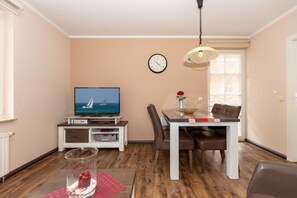 Una sala de estar con televisor, mesa de comedor, sillas, un reloj y un florero con una flor.