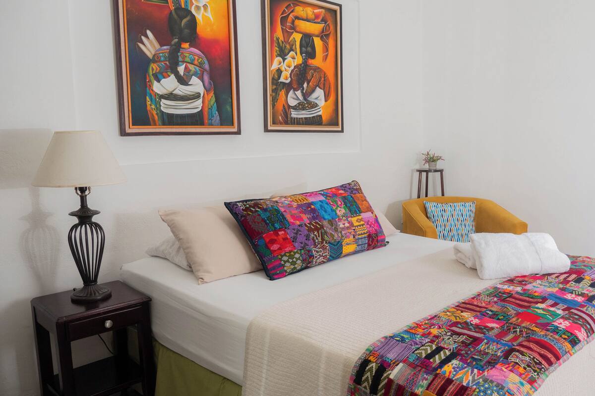 Habitación de hotel con cama, mesita de noche, lámpara, dos cuadros, una silla y una ventana con cortinas.