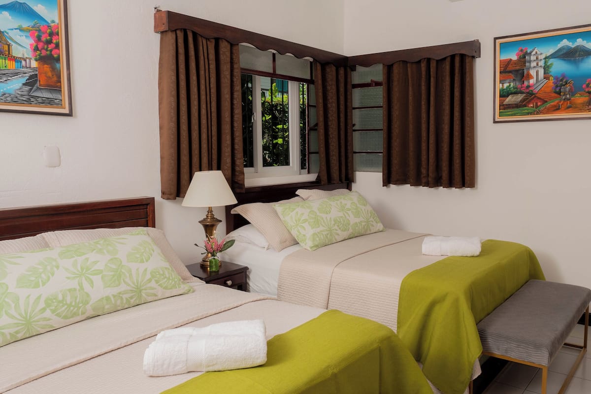 Habitación de hotel con dos camas, una mesita de noche con lámpara, un banco y dos cuadros en la pared.