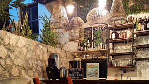 Un área de bar al aire libre con luces colgantes de mimbre, una pared de piedra y una variedad de botellas y vasos en estanterías.