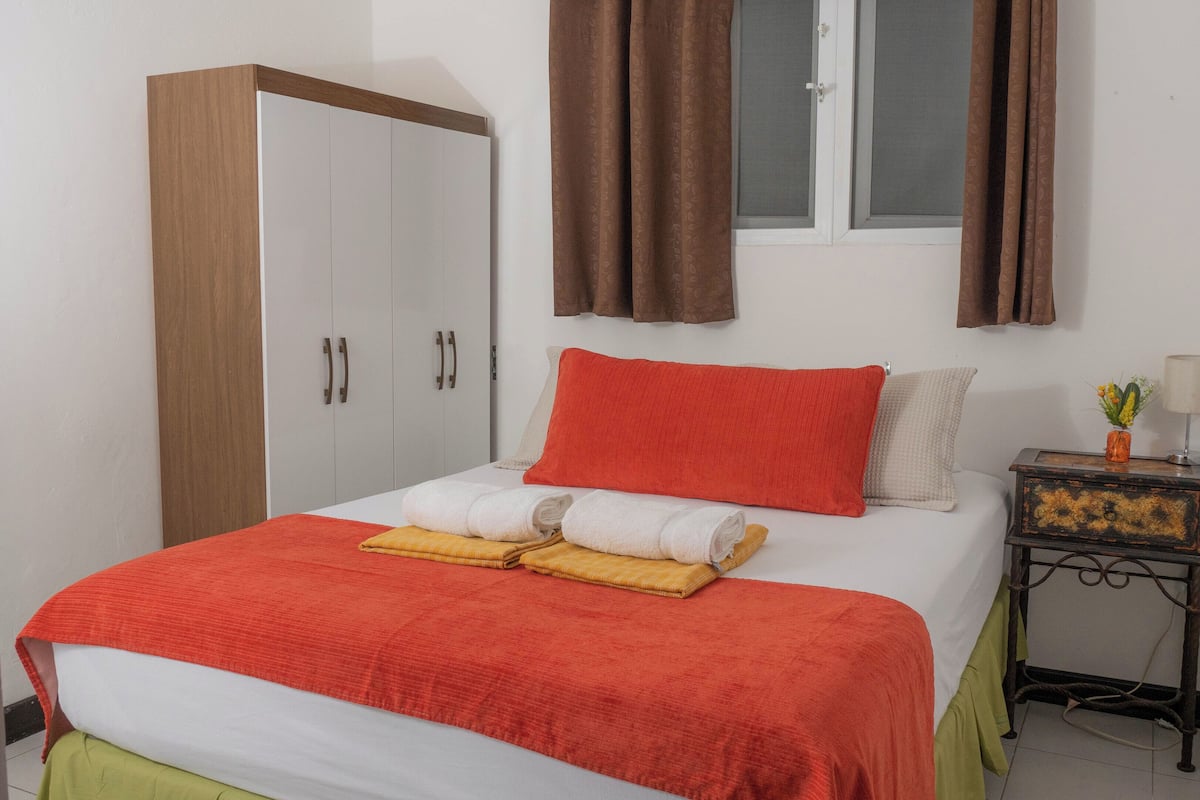 Un dormitorio con cama, armario, ventana con cortinas, mesita de noche con lámpara y una planta pequeña.