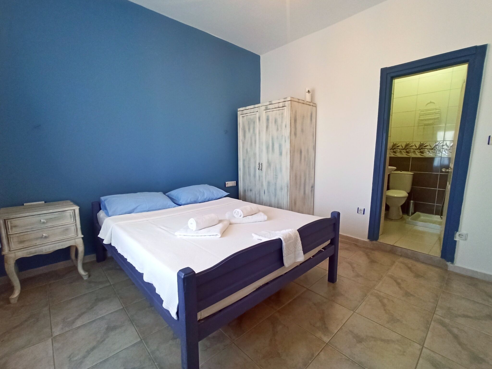 A bedroom with a bed, a nightstand, a wardrobe, and a bathroom visible through an open door.