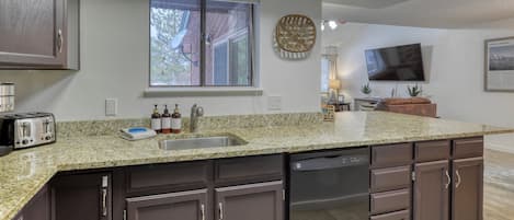A kitchen with a granite countertop, dark cabinetry, a dishwasher, and a window with blinds.