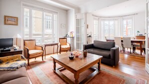 A spacious living room with a wooden coffee table, a sofa, and a television. There are two chairs, a dining table, and a view of the city through large windows. - Flowers from Paris, by Veeve (Paris)