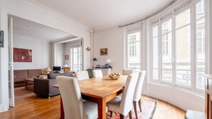 A spacious living area with a wooden dining table, white chairs, a sofa, and a coffee table. There are large windows providing natural light and a view of the outside. - Flowers from Paris, by Veeve (Paris)
