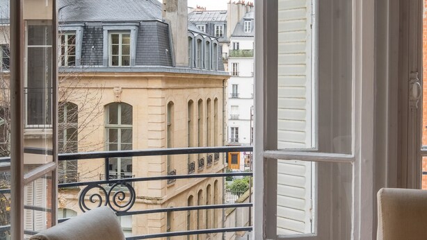 A room with a wooden table and chairs, a large window with shutters, and a view of neighboring buildings. - Flowers from Paris, by Veeve (Paris)