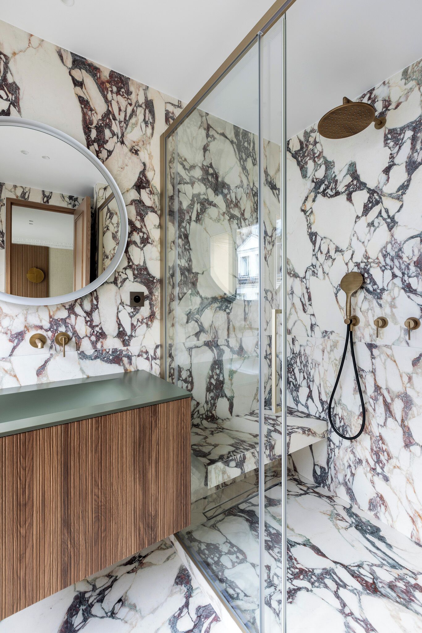 Un baño con pared de mármol, tocador de madera, cabina de ducha de vidrio y espejo redondo.