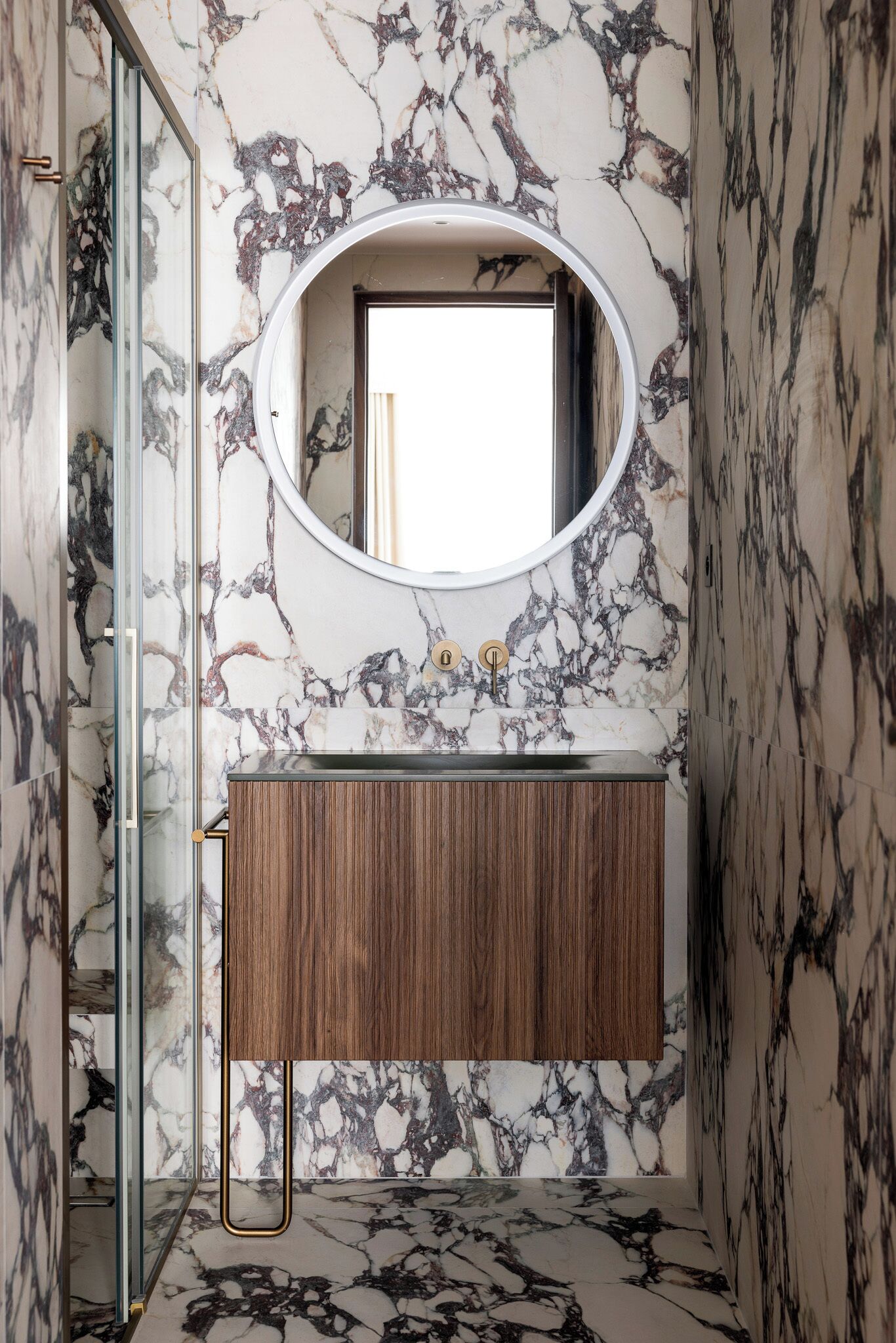 Un baño con pared de mármol, tocador de madera, espejo redondo y cabina de ducha de vidrio.