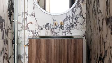 Un baño con pared de mármol, tocador de madera, espejo redondo y cabina de ducha de vidrio.