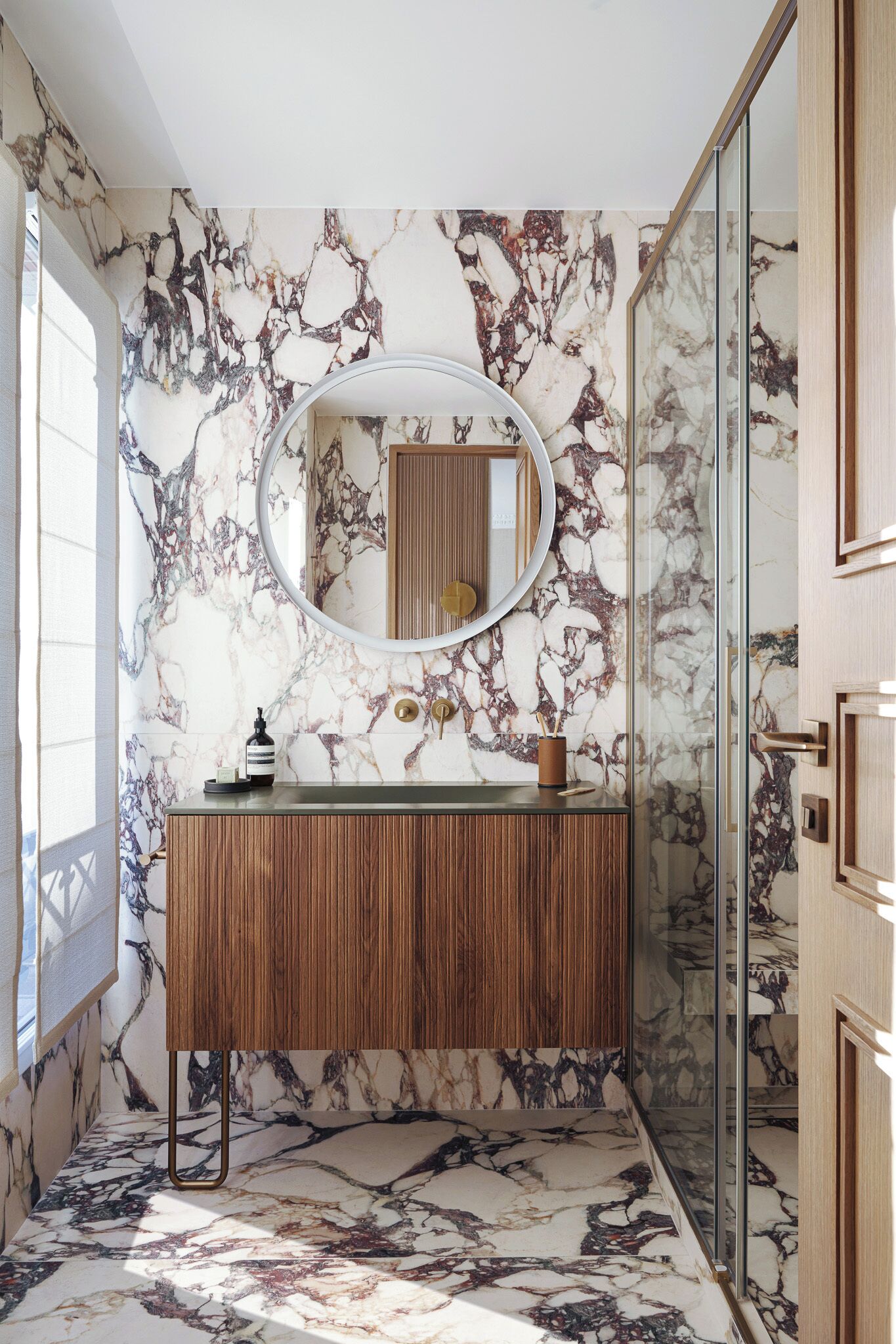 A bathroom with a marble wall, a wooden vanity, a round mirror, and a glass shower enclosure.
