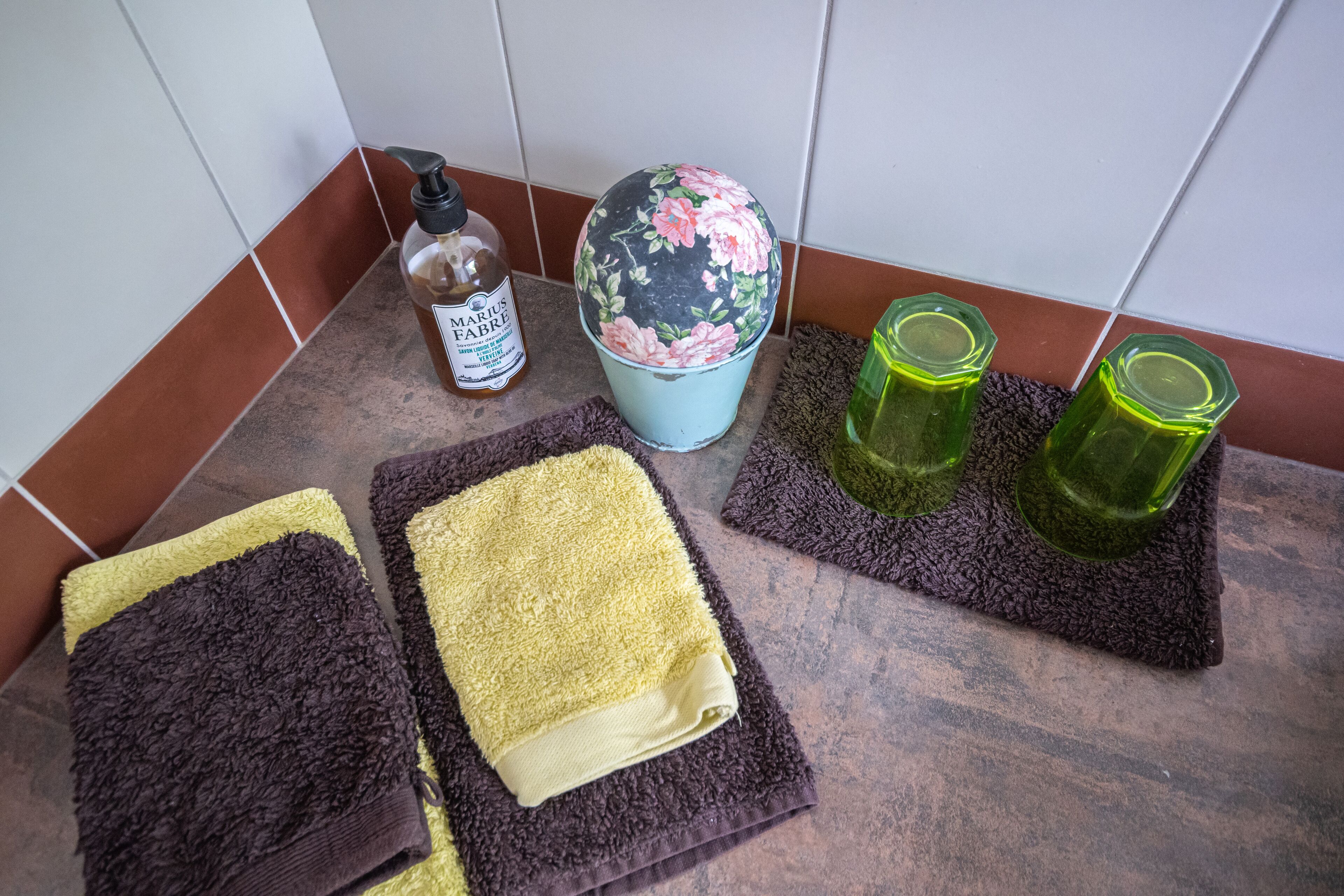 Un baño con piso de baldosas, una botella de jabón para manos, un cubo estampado con motivos florales y dos toallas sobre un tapete de baño.