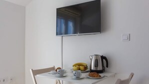 A small dining area with a white table, two chairs, a TV mounted on the wall, a kettle, a plate with bananas, a cup, and a croissant. - Studio Apartman Blue View (Dubrovnik)