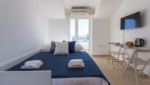 A hotel room with a bed, a desk with a chair, a flat-screen TV, and a view of the sea through a large window. - Studio Apartman Blue View (Dubrovnik)