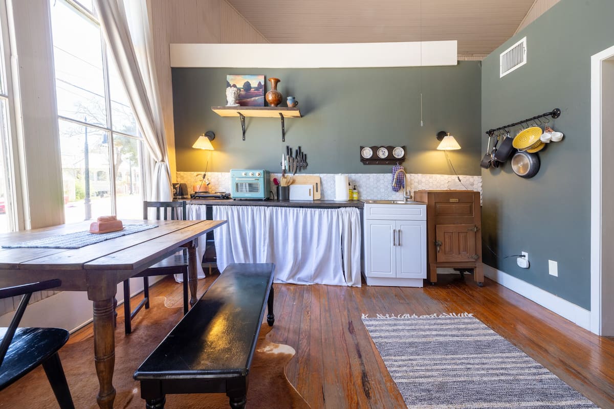 A kitchen with wooden flooring, a dining table, and a bench. There is a window with curtains, a wall-mounted shelf with decorative items, and a wall-mounted pot rack.