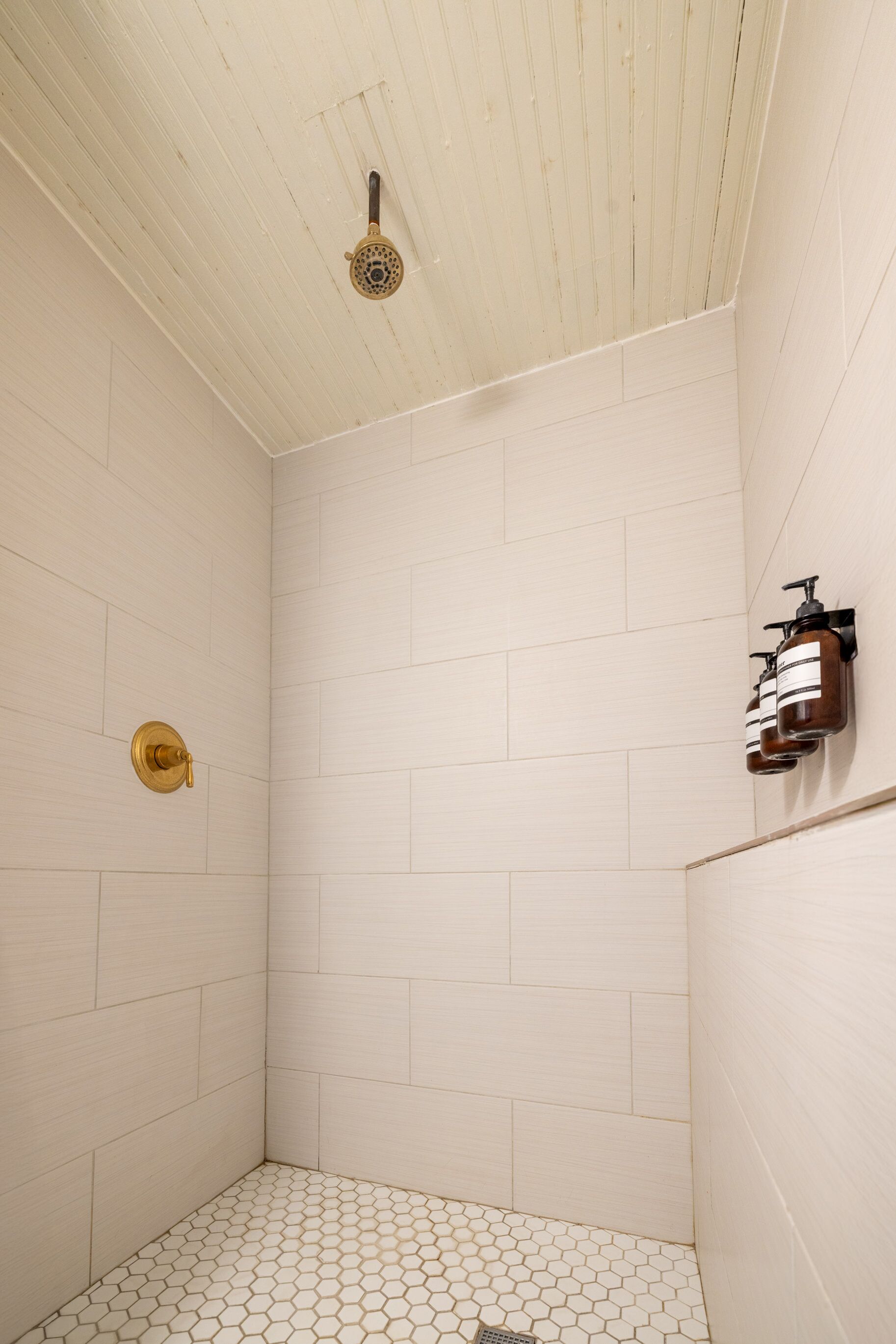 Un baño pequeño con azulejos blancos, suelo de baldosas hexagonales, cabezal de ducha dorado y dispensador de jabón en la pared.