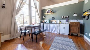 A spacious kitchen with wooden floors, a dining area with a bench, and a view through large windows. - The Downtown Abbey (San Marcos)