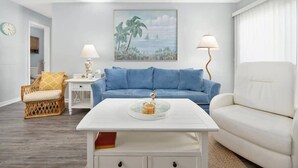 A living room with a white coffee table, a blue sofa, a white sofa, a wicker chair, and a painting on the wall.