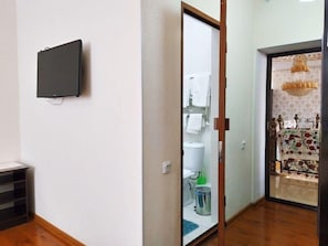 A hotel room with a television mounted on the wall, a wooden floor, a door leading to a bathroom with a toilet and sink, and a patterned curtain. - Euro-asia (Khiva)