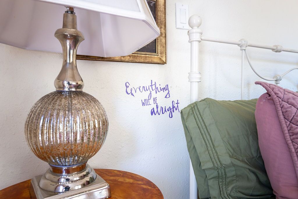 Een lamp op een houten tafel, een bed met een wit metalen frame en een muur met een inspirerende quote.