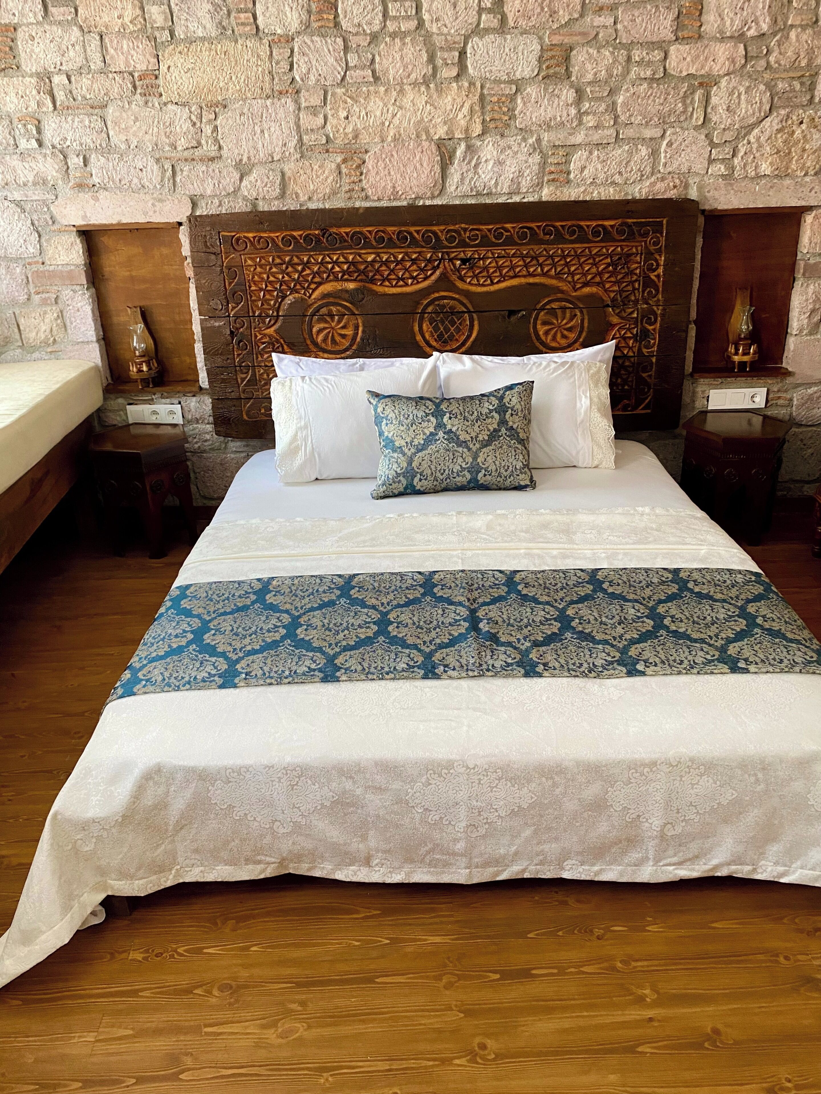 Ein Bett mit einem gemusterten Überwurf, einem Kopfstück mit aufwendigen Schnitzereien und einer dahinterliegenden Steinwand.