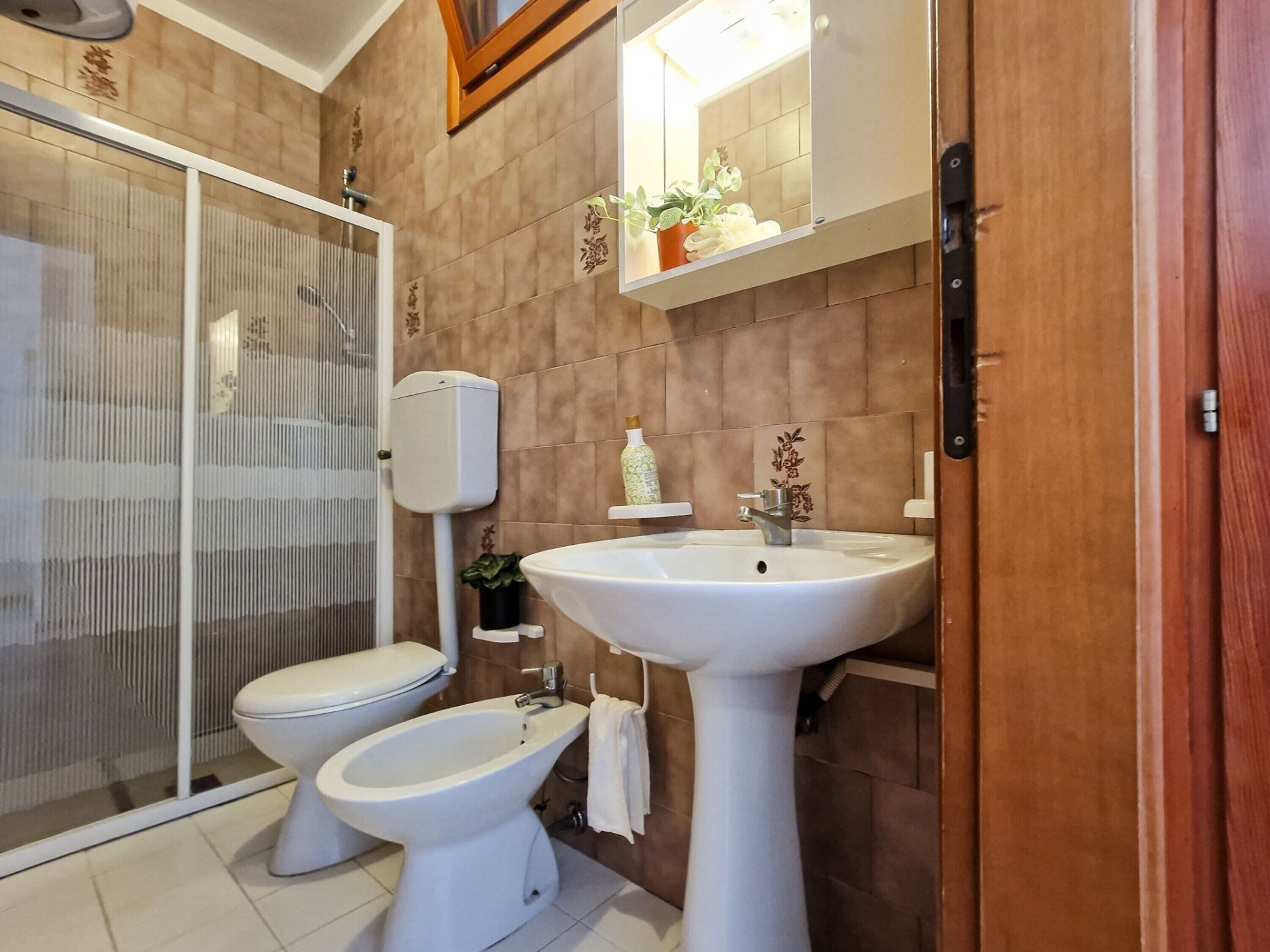 Une salle de bain comprenant des toilettes, un bidet et un lavabo, une douche avec une porte en verre et une porte en bois.