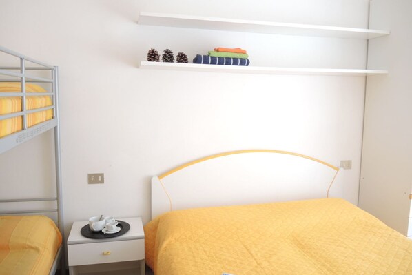 A room with a single bed, a nightstand with a cup and saucer, a small shelf with decorative items, and a bunk bed in the background. - Beach Retreat for up to 5 Guests-beahost (San Michele al Tagliamento)