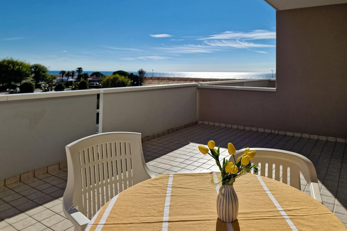 A balcony with a table and chairs, a vase with yellow flowers, and a view of the sea.