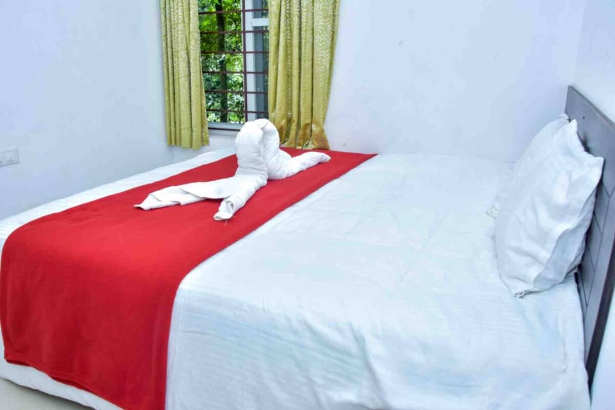 A neatly made bed with a red blanket and white linens, a window with green curtains, and a view of greenery outside.