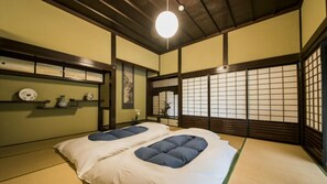 A traditional Japanese room with tatami flooring, a low wooden bed with cushions, a shoji screen door, and a hanging light fixture. - Kondoke Inn (Toon)