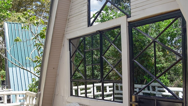 Una terraza de madera con sillas de mimbre blancas y una mesa, una puerta negra y un ventanal con diseño de rombos.