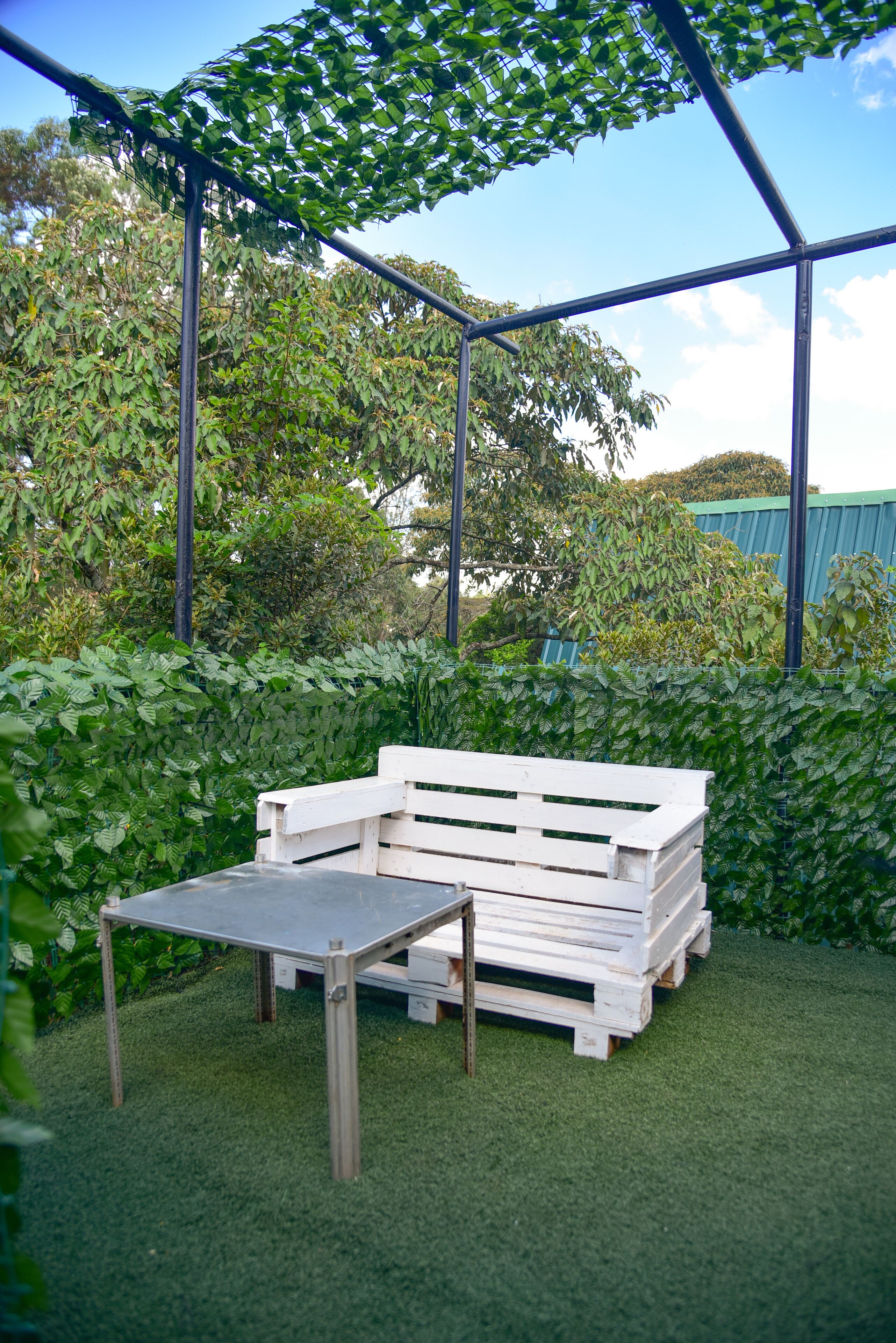 Conjunto de banco e mesa de madeira branca sobre grama sintética, cercado por folhagem verde exuberante e uma estrutura metálica acima.