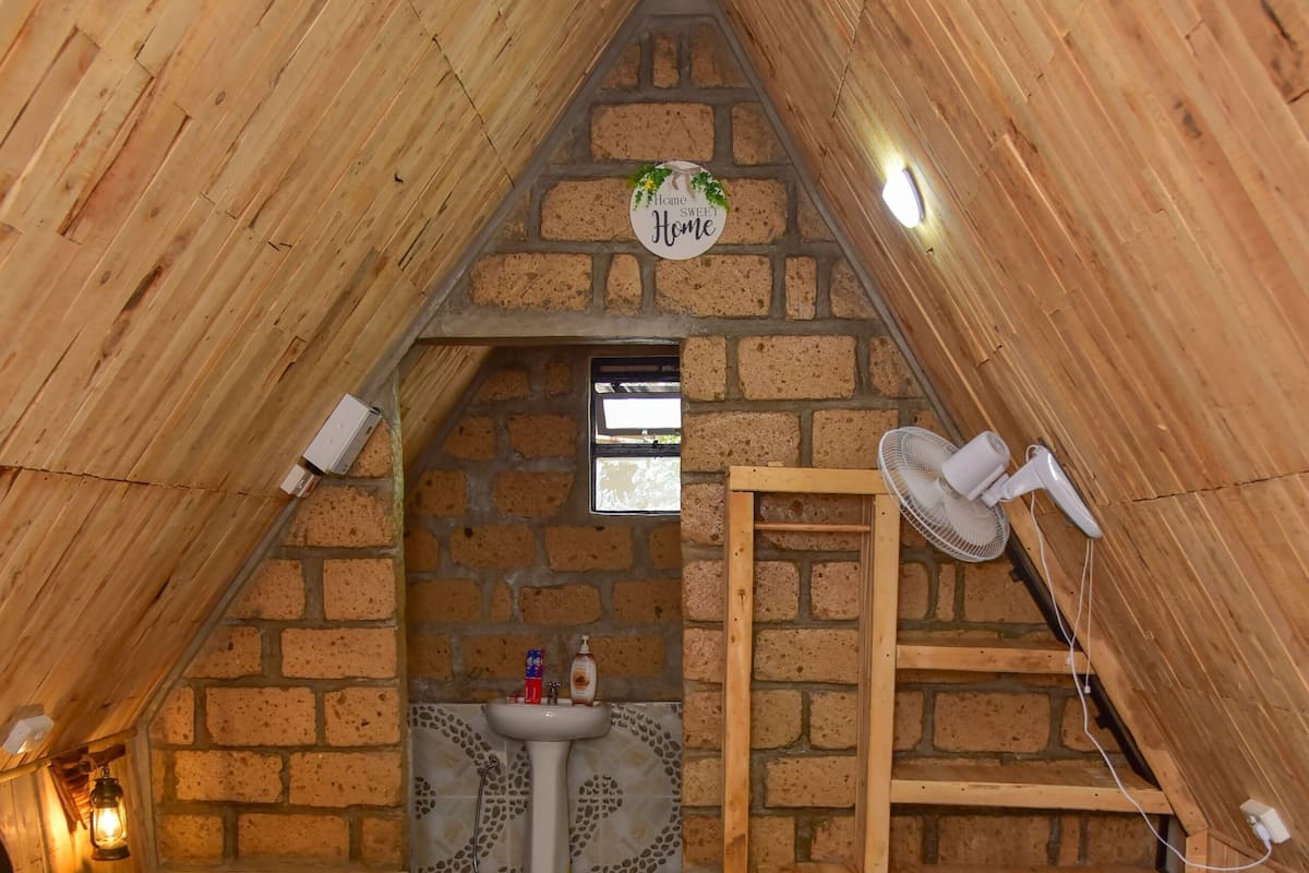 A cozy attic bedroom with a wooden sloped ceiling, a brick wall, a small window, a wooden ladder, a bedside table with a lamp, and a neatly made bed with a towel.