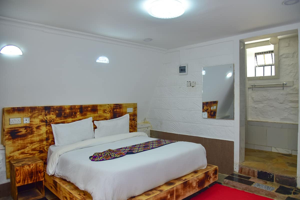 A bedroom with a wooden headboard, white bedding, a red carpet, a small bedside table, a mirror, and a shower area.