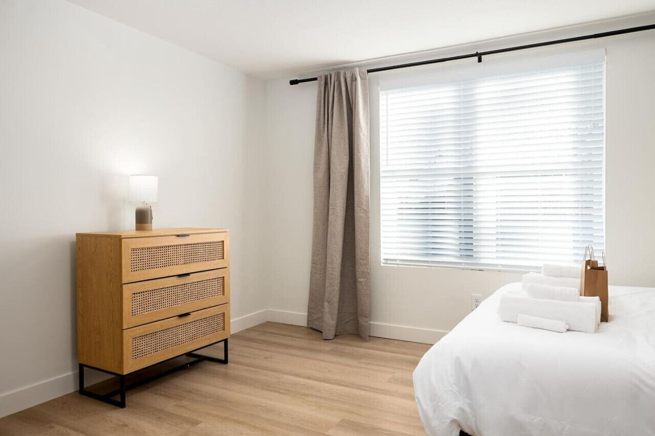 Ett hotellrum med en byrå i trä, en säng med vita sängkläder, ett fönster med persienner och en gardin.