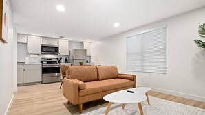 Ein modernes Wohnzimmer mit einer Couch und einem Couchtisch, eine K üche mit weißen Schränken und Geräten sowie ein großes Fenster mit Jalousien.