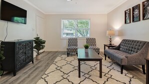 A living room with a ceiling fan, a TV mounted on the wall, two patterned armchairs, a sofa, a coffee table, and a window with blinds. - CorporateHousingSavannah.com (Savannah)